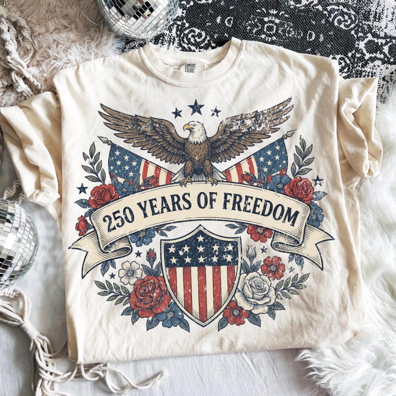 May include: Cream-colored t-shirt featuring a patriotic design. The graphic includes an eagle with outstretched wings, American flags, a shield, and the text "250 YEARS OF FREEDOM" on a banner. Floral elements surround the design.