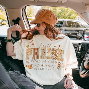 May include: A cream-coloured t-shirt featuring the text "I will PRAISE Him in every season" in a mix of patterned and floral fonts. The shirt has a crew neck and short sleeves. The design includes autumn-themed leaves and Psalm 146:2.