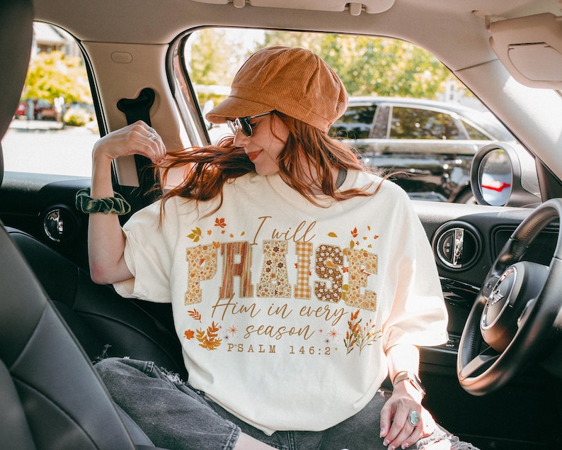 May include: A cream-coloured t-shirt featuring the text "I will PRAISE Him in every season" in a mix of patterned and floral fonts. The shirt has a crew neck and short sleeves. The design includes autumn-themed leaves and Psalm 146:2.
