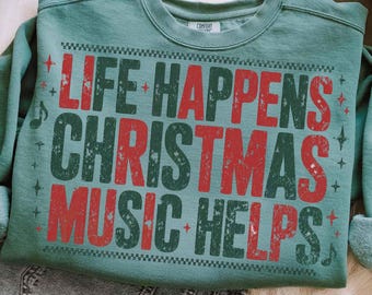 May include: A teal green sweatshirt with the text "LIFE HAPPENS CHRISTMAS MUSIC HELPS" in distressed red and green lettering. The design includes musical notes and star accents, perfect for the holiday season.