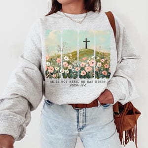May include: A gray sweatshirt with a floral design and a cross on a hill. The text "He is not here. He has risen. Matthew 28:6" is printed on the sweatshirt.