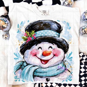 May include: A white t-shirt featuring a graphic illustration of a smiling snowman wearing a black top hat with a light blue polka dot band, a light blue scarf, and a sprig of holly with red berries. The snowman has rosy cheeks and an orange nose.
