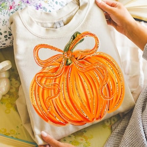 May include: A beige sweatshirt featuring a large orange pumpkin design with gold glitter accents. The pumpkin has a green stem and a decorative bow. The sweatshirt is folded and held by a person.