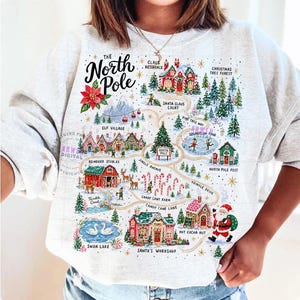 May include: A light gray sweatshirt featuring a colorful graphic map of the North Pole. The design includes illustrations of Santa's workshop, reindeer stables, elf village, and various holiday landmarks with text labels for each location.
