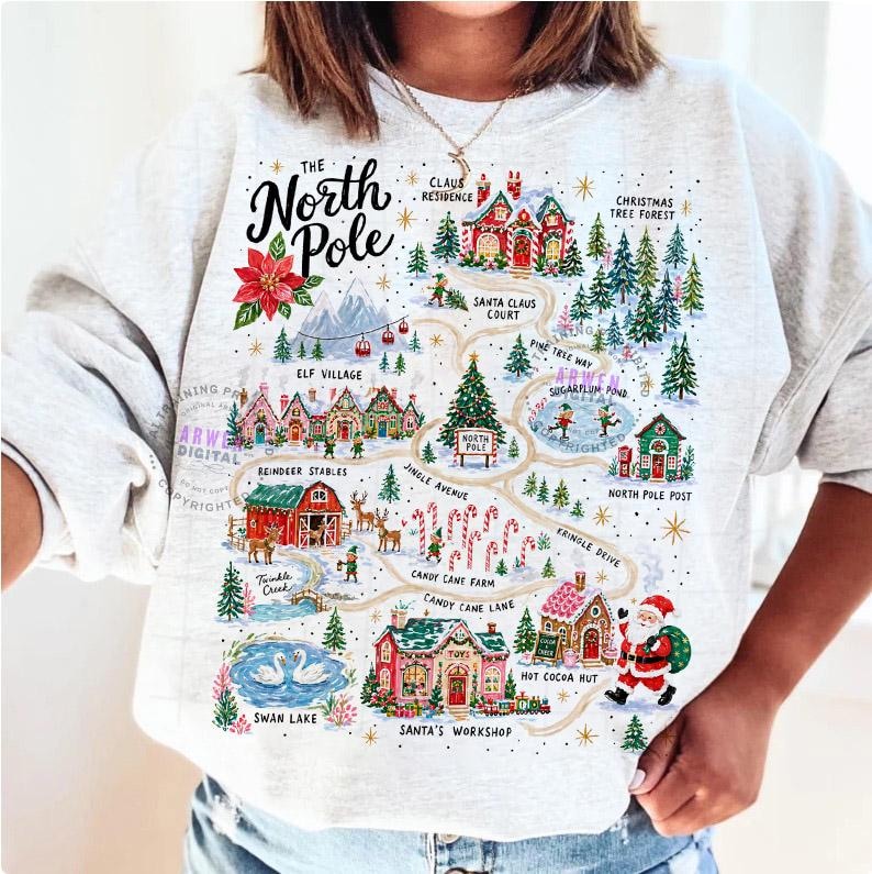 May include: A light gray sweatshirt featuring a colorful graphic map of the North Pole. The design includes illustrations of Santa's workshop, reindeer stables, elf village, and various holiday landmarks with text labels for each location.