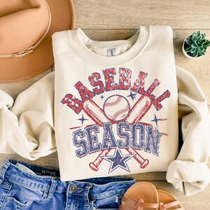 May include: A cream-colored sweatshirt with a vintage-style graphic of crossed baseball bats and a baseball with the text "Baseball Season" in red, blue, and white.