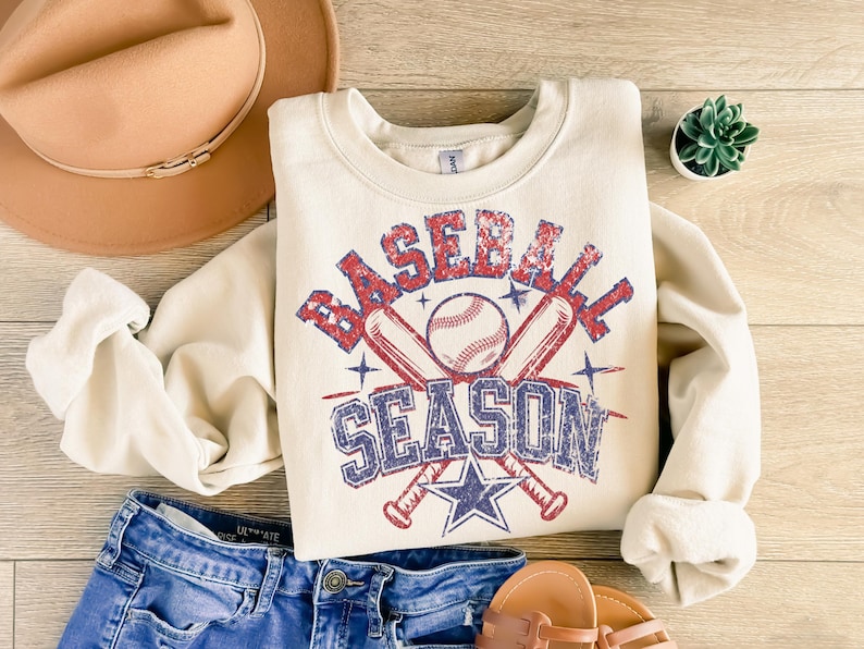 May include: A cream-colored sweatshirt with a vintage-style graphic of crossed baseball bats and a baseball with the text "Baseball Season" in red, blue, and white.