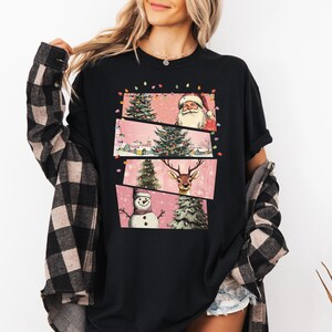 May include: Black t-shirt featuring a festive Christmas design. The graphic includes Santa Claus, a reindeer, a snowman, and Christmas trees. The design is set against a pink background with string lights.