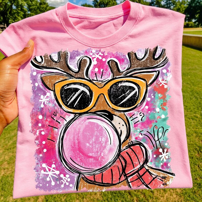 Op de afbeelding: Roze T-shirt met een cartoon rendier met een zonnebril die een kauwgom bel blaast. Het rendier heeft bruine geweien en een rood-zwarte geruite sjaal. De achtergrond heeft roze, paarse en turquoise accenten met witte sneeuwvlokken.