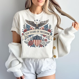 May include: Cream-colored t-shirt featuring a patriotic design. The graphic includes an eagle, American flags, a shield, and the text "250 YEARS OF FREEDOM" on a banner. Floral elements surround the central design.