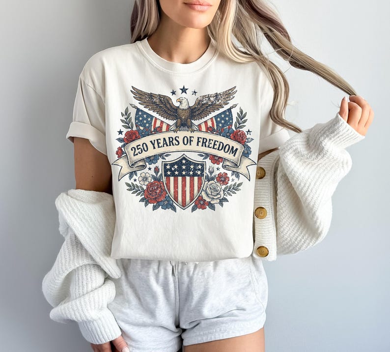 May include: Cream-colored t-shirt featuring a patriotic design. The graphic includes an eagle, American flags, a shield, and the text "250 YEARS OF FREEDOM" on a banner. Floral elements surround the central design.