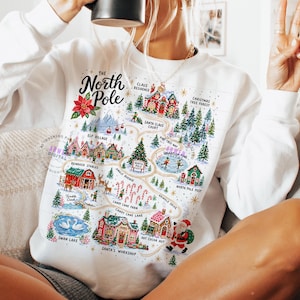 May include: A white long-sleeve sweatshirt featuring a colorful graphic map of the North Pole. The design includes illustrations of Santa's workshop, reindeer stables, candy cane farm, and various festive buildings with text labels for each location.