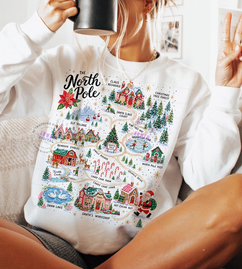 May include: A white long-sleeve sweatshirt featuring a colorful graphic map of the North Pole. The design includes illustrations of Santa's workshop, reindeer stables, candy cane farm, and various festive buildings with text labels for each location.