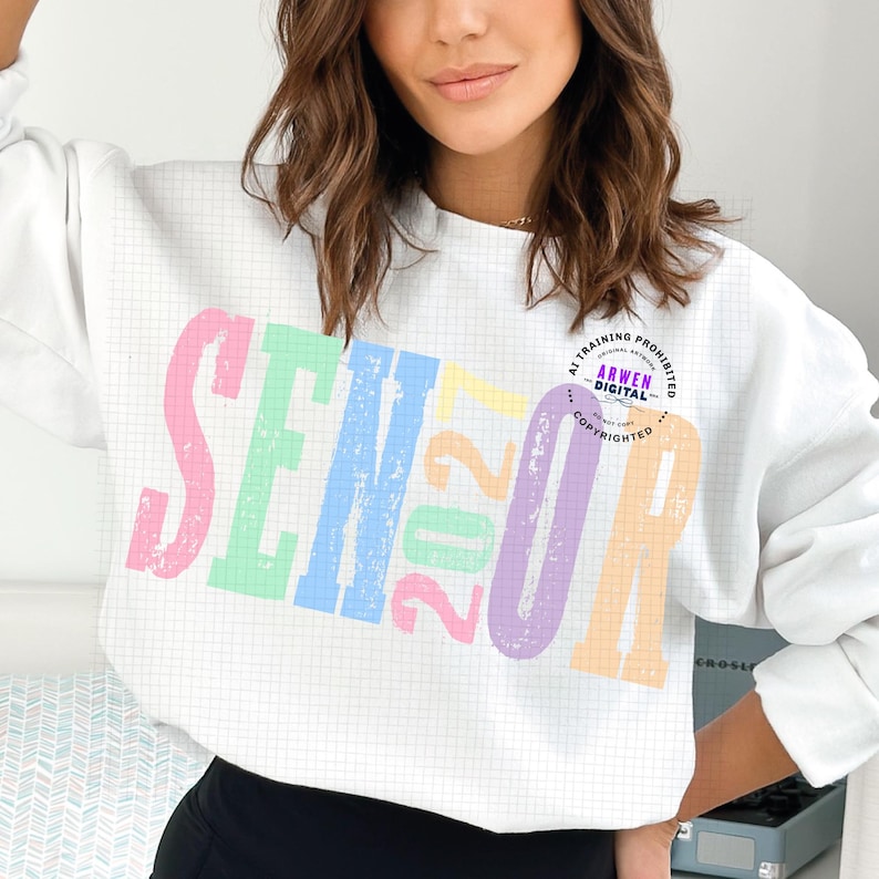 Puede incluir: Una sudadera blanca de manga larga con un fondo de patr&oacute;n de cuadr&iacute;cula y la palabra SENIOR en letras grandes, desgastadas y de colores pastel. El n&uacute;mero 2027 est&aacute; impreso en el centro en colores amarillo, rosa y verde pastel.