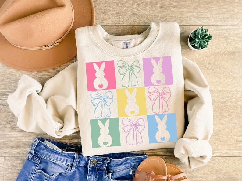 May include: A cream-colored sweatshirt with a graphic design of pastel-colored bunnies and bows. The design is in a grid pattern with each bunny and bow in a different color.