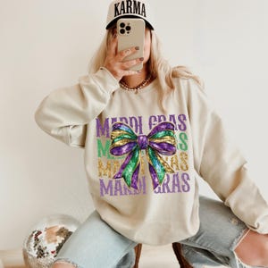 Puede incluir: Una sudadera beige con un lazo de purpurina morado, verde y dorado y el texto "Mardi Gras" repetido varias veces.