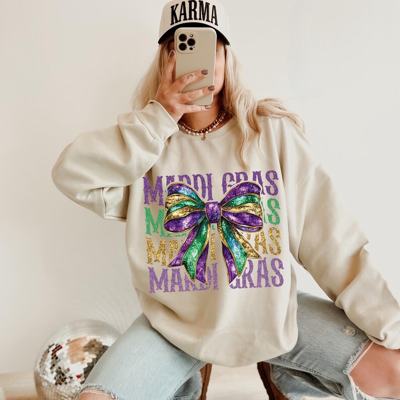 Puede incluir: Una sudadera beige con un lazo de purpurina morado, verde y dorado y el texto "Mardi Gras" repetido varias veces.