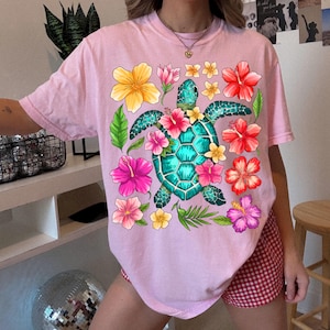 May include: Pink t-shirt featuring a graphic of a green sea turtle surrounded by colourful flowers. The turtle has a blue shell and is surrounded by pink, yellow and red flowers.