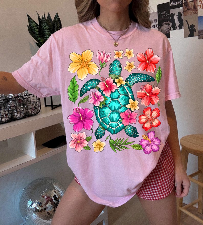 May include: Pink t-shirt featuring a graphic of a green sea turtle surrounded by colourful flowers. The turtle has a blue shell and is surrounded by pink, yellow and red flowers.