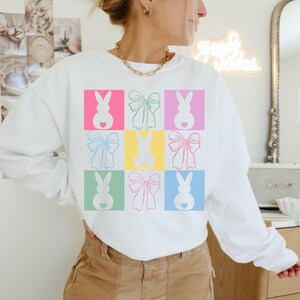 May include: White sweatshirt with a colorful graphic design of bunnies and bows. The design is repeated in a grid pattern.