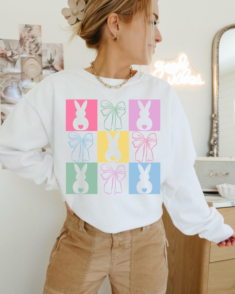 May include: White sweatshirt with a colorful graphic design of bunnies and bows. The design is repeated in a grid pattern.
