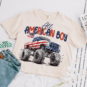 May include: Cream-colored t-shirt featuring a graphic of a monster truck with an American flag design and the text "All AMERICAN BOY" in red and blue. The shirt is paired with denim jeans and a green and white hair clip.