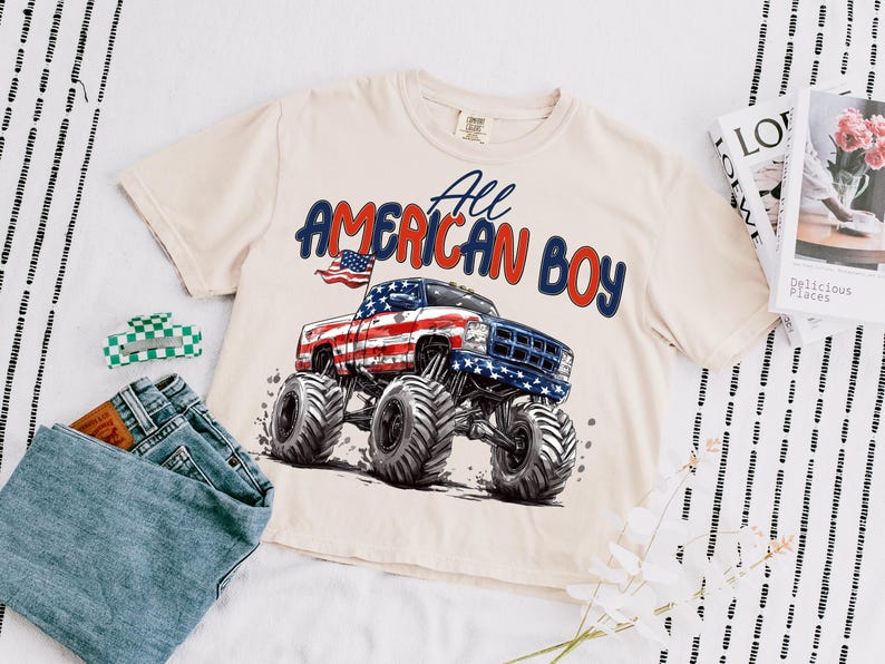 May include: Cream-colored t-shirt featuring a graphic of a monster truck with an American flag design and the text "All AMERICAN BOY" in red and blue. The shirt is paired with denim jeans and a green and white hair clip.