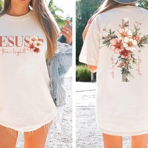 May include: A white t-shirt with a floral cross design on the back and the text "Jesus is the light" and a floral design on the front.