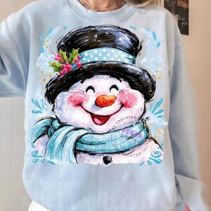 May include: A light blue sweatshirt featuring a graphic illustration of a smiling snowman wearing a black top hat with a light blue polka dot band, a light blue scarf, and a sprig of holly with red berries. The snowman has a carrot nose.