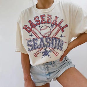 May include: A white t-shirt with a vintage baseball graphic. The graphic features two crossed baseball bats, a baseball, and the text "Baseball Season".