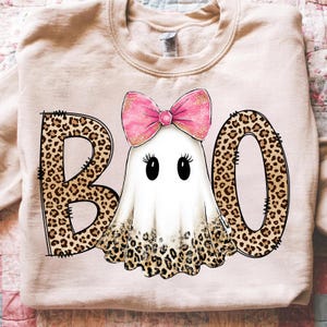 May include: A light beige sweatshirt with a Halloween-themed design. The word "BOO" is spelled out with leopard print letters. A cartoon ghost with a pink bow is in the center. The ghost has black eyes and eyelashes.