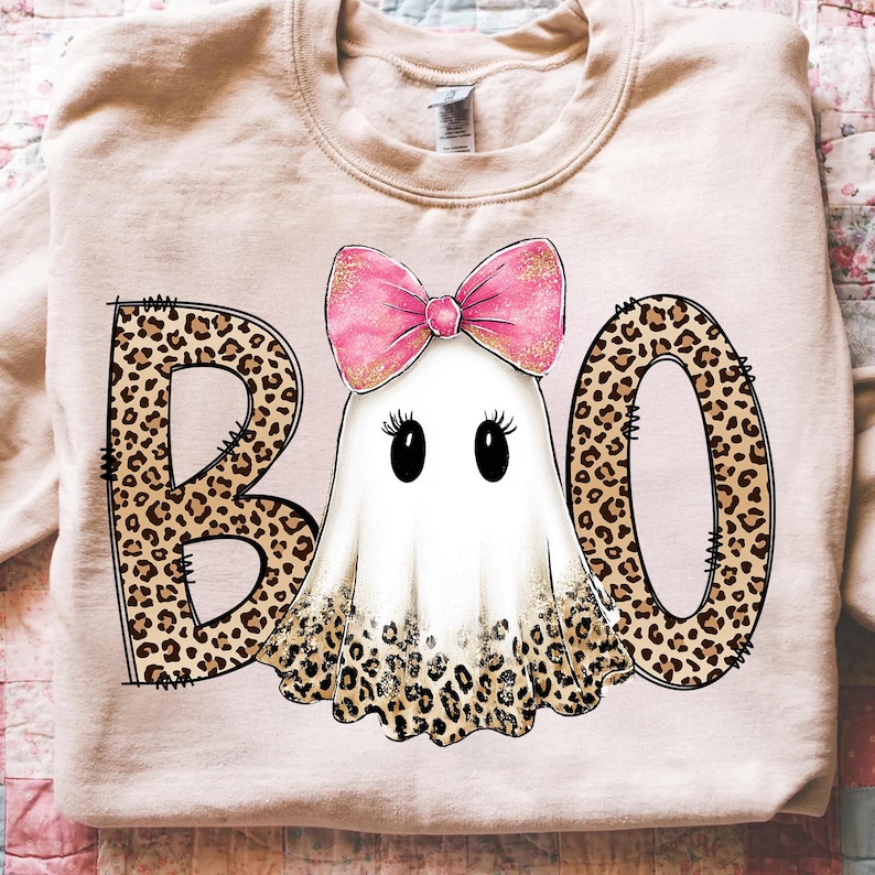 May include: A light beige sweatshirt with a Halloween-themed design. The word "BOO" is spelled out with leopard print letters. A cartoon ghost with a pink bow is in the center. The ghost has black eyes and eyelashes.