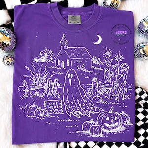 May include: A purple t-shirt featuring a white graphic print of a ghost, a church, pumpkins, and corn stalks. The text "LIVE HERE NOW" is printed on a small sign near the ghost. The shirt has a crew neckline and a Comfort Colors brand label.