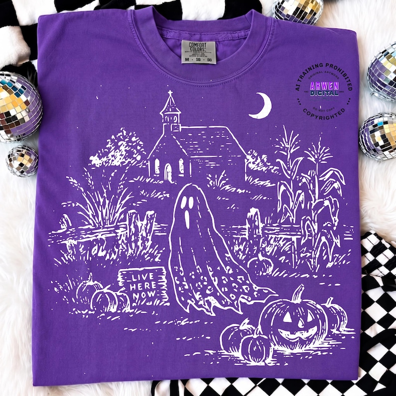 May include: A purple t-shirt featuring a white graphic print of a ghost, a church, pumpkins, and corn stalks. The text "LIVE HERE NOW" is printed on a small sign near the ghost. The shirt has a crew neckline and a Comfort Colors brand label.