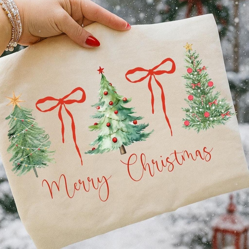 May include: A beige tote bag featuring watercolor illustrations of Christmas trees and red bows. The words "Merry Christmas" are written in red script below the trees. The bag is held by a person.