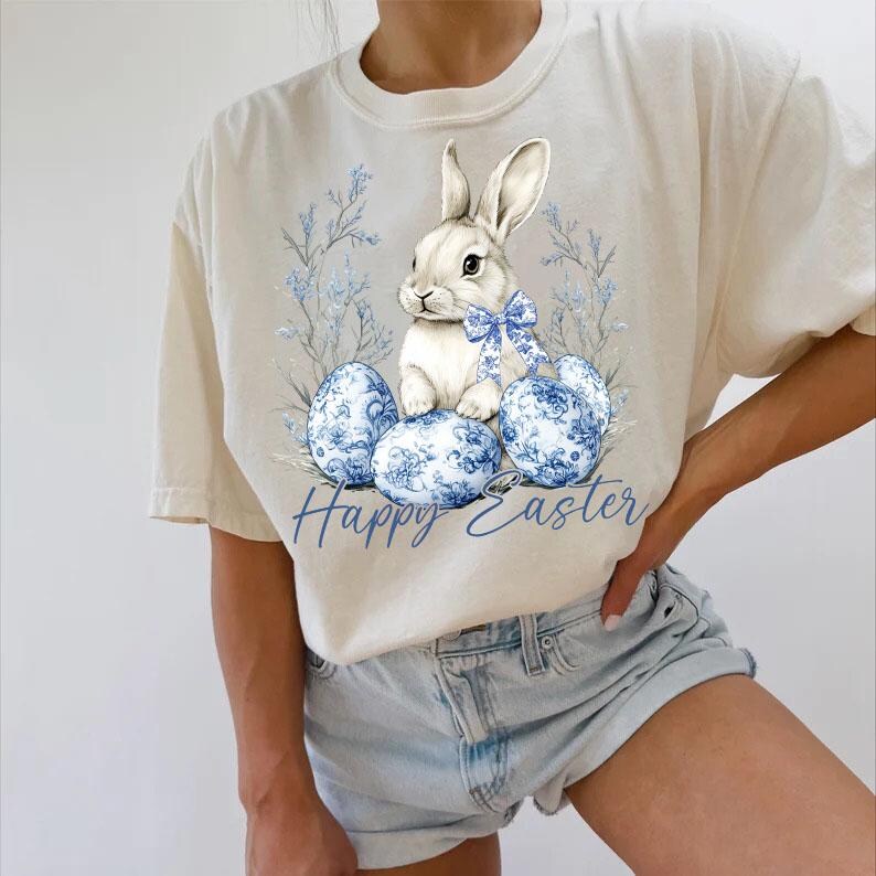 May include: Cream-colored t-shirt featuring a white bunny with a blue bow, surrounded by blue floral Easter eggs and foliage. The words "Happy Easter" are written in blue script below the design. The shirt has short sleeves.