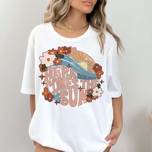 May include: White t-shirt with a retro-style graphic design featuring a sun, waves, and flowers. The text "Here Comes The Sun" is printed in a vintage font.