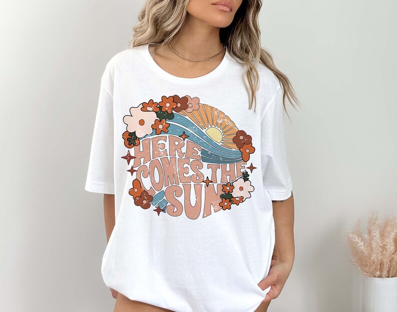 May include: White t-shirt with a retro-style graphic design featuring a sun, waves, and flowers. The text "Here Comes The Sun" is printed in a vintage font.