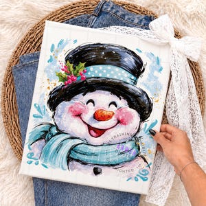 May include: A square canvas print featuring a cheerful snowman illustration with a black top hat, a light blue scarf, and pink cheeks. The snowman is accented with holly berries and blue paint splatters. The artwork has a white grid background.