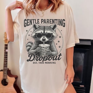 Puede incluir: Camiseta color crema con un mapache con gafas de sol y sosteniendo una bebida. La camiseta tiene el texto "GENTLE PARENTING" encima del mapache y "Dropout EST. THIS MORNING" debajo. El dise&ntilde;o es en negro y gris.