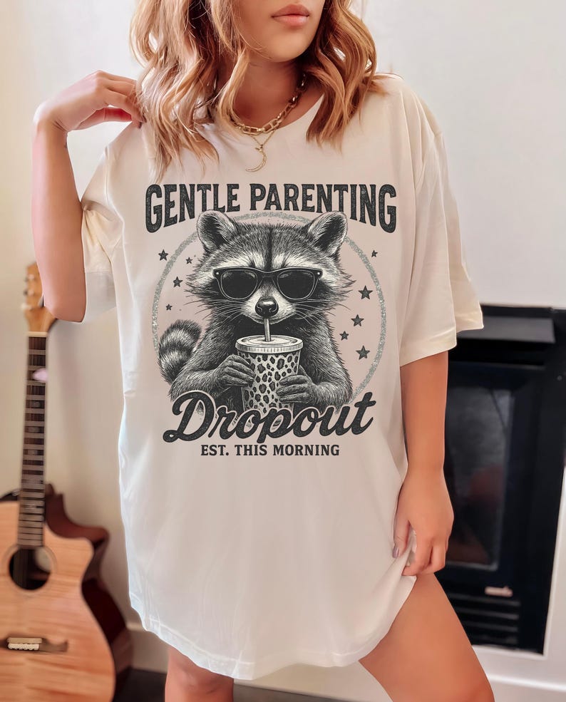 Puede incluir: Camiseta color crema con un mapache con gafas de sol y sosteniendo una bebida. La camiseta tiene el texto "GENTLE PARENTING" encima del mapache y "Dropout EST. THIS MORNING" debajo. El dise&ntilde;o es en negro y gris.