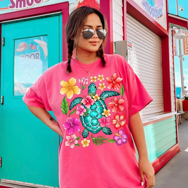 May include: Pink t-shirt featuring a colourful graphic of a sea turtle surrounded by tropical flowers. The turtle is green and blue with a detailed shell.