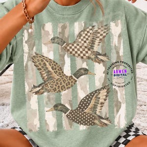 Peut inclure: Un sweat-shirt à col rond vert sauge orné d'un motif de trois canards en vol sur un fond à rayures verticales délavées. Le canard du haut a un motif à carreaux, celui du milieu un imprimé léopard et celui du bas un motif à pois.