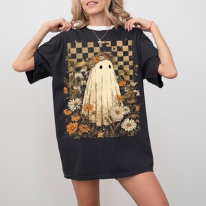 Op de afbeelding: Zwart T-shirt met een spookafbeelding, omringd door bloemen en tegen een geblokte achtergrond. Het ontwerp gebruikt beige, oranje en wit, wat een vintage esthetiek cre&euml;ert. Het T-shirt heeft een comfortabele pasvorm.