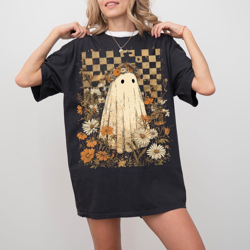 Op de afbeelding: Zwart T-shirt met een spookafbeelding, omringd door bloemen en tegen een geblokte achtergrond. Het ontwerp gebruikt beige, oranje en wit, wat een vintage esthetiek cre&euml;ert. Het T-shirt heeft een comfortabele pasvorm.