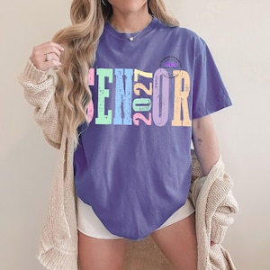 Puede incluir: Una camiseta morada de manga corta con el texto SENIOR 2027 en una fuente desgastada y multicolor, con letras en tonos rosa, verde, azul y amarillo. La camiseta se lleva con un c&aacute;rdigan de punto beige abierto y pantalones cortos blancos.