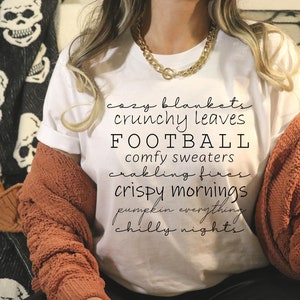 May include: A white t-shirt with the word "FOOTBALL" in large black letters. The shirt also features the phrases "cozy blankets," "crunchy leaves," "comfy sweaters," "crispy mornings," and "chilly nights" in a script font. A woman is wearing the shirt with a gold chain necklace and a rust-colored sweater.