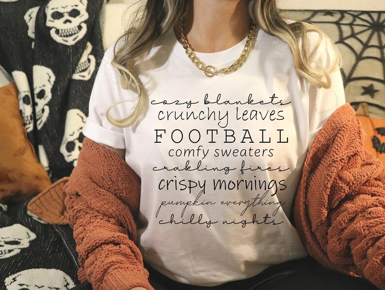 May include: A white t-shirt with the word "FOOTBALL" in large black letters. The shirt also features the phrases "cozy blankets," "crunchy leaves," "comfy sweaters," "crispy mornings," and "chilly nights" in a script font. A woman is wearing the shirt with a gold chain necklace and a rust-colored sweater.