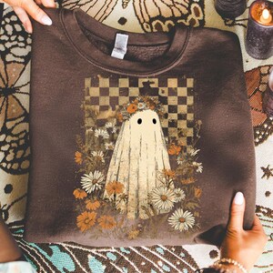 Op de afbeelding: Een bruine sweatshirt met een grillig spookontwerp. Het spook is gehuld in een wit laken, versierd met een bloemenkrans en geplaatst tegen een geblokte achtergrond. Het ontwerp is omgeven door madeliefjes en andere bloemen.