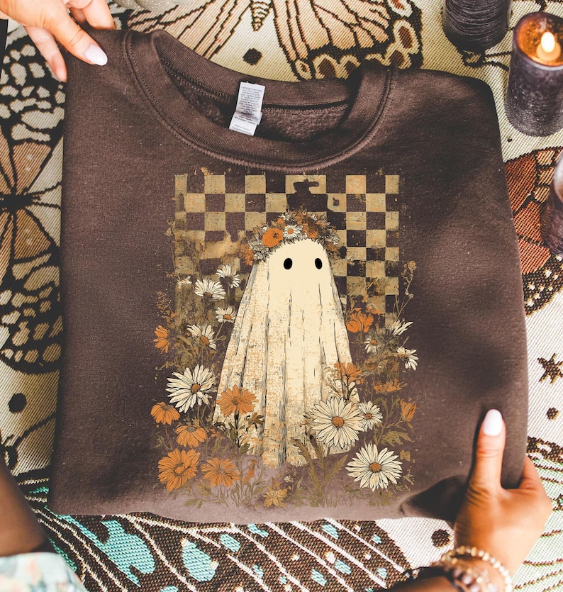 Op de afbeelding: Een bruine sweatshirt met een grillig spookontwerp. Het spook is gehuld in een wit laken, versierd met een bloemenkrans en geplaatst tegen een geblokte achtergrond. Het ontwerp is omgeven door madeliefjes en andere bloemen.